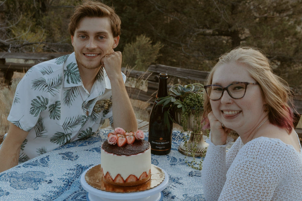 Newly weds sitting at a picnic table with a round blue and white table cloth. On the table is a strawberry cake, bottle of champagne and green plant filled bouquet.