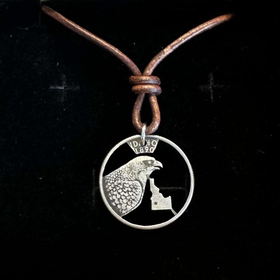 Idaho State Quarter Hand Cut Coin Necklace with a leather cord in a brown kraft box on a black velvet background. - Mountainstruck Coin Jewelry