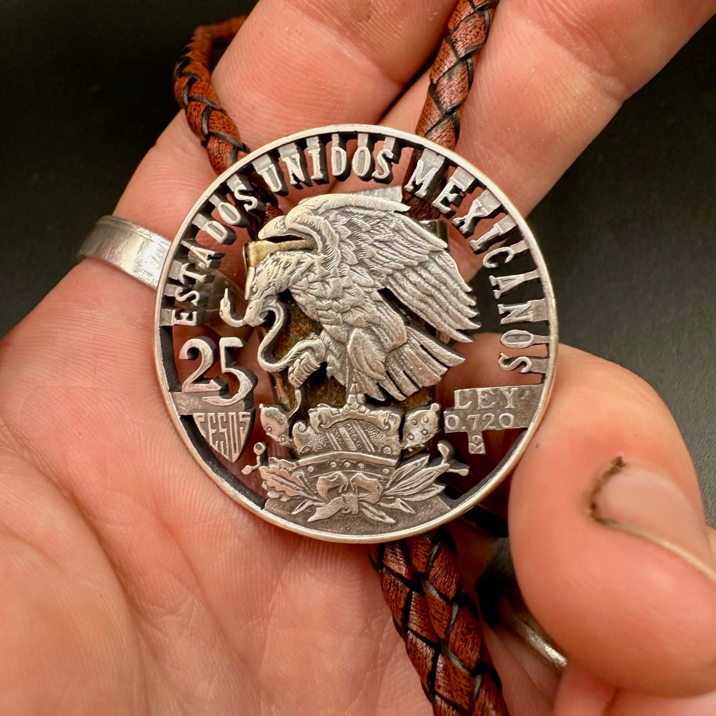 Mexico Silver 25 Pesos Hand Cut Coin Bolo Tie with darker brown leather cord on a hand for display. - Mountainstruck Coin Jewelry