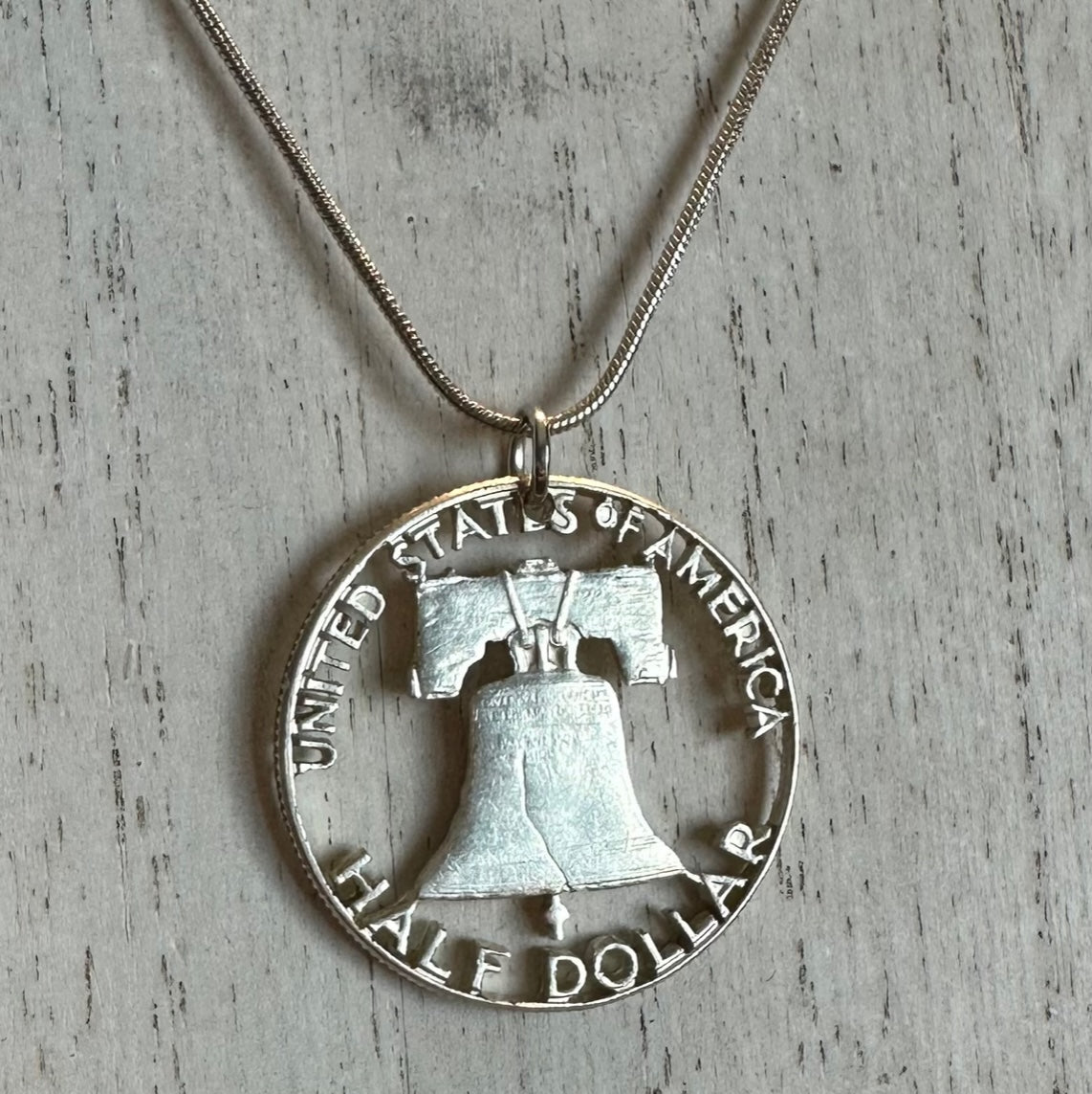Cracked Liberty Bell U.S. Half Dollar Hand Cut Coin Necklace with snake chain on a white wood background. - Mountainstruck Coin Jewelry