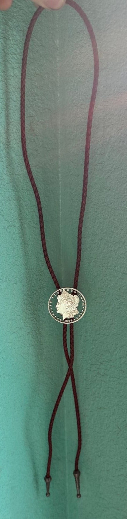 U.S. Morgan Hand Cut Silver Dollar Coin Bolo Tie (front) hanging in front of teal background. - Mountainstruck Coin Jewelry
