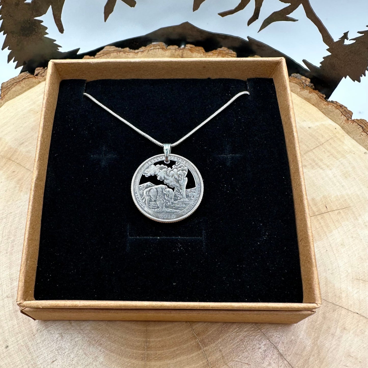 U.S. Yellowstone National Park Quarter Hand Cut Coin Necklace in a brown kraft box on a black velvet background on top of a light wood round with a metal mountain and tree behind. - Mountainstruck Coin Jewelry