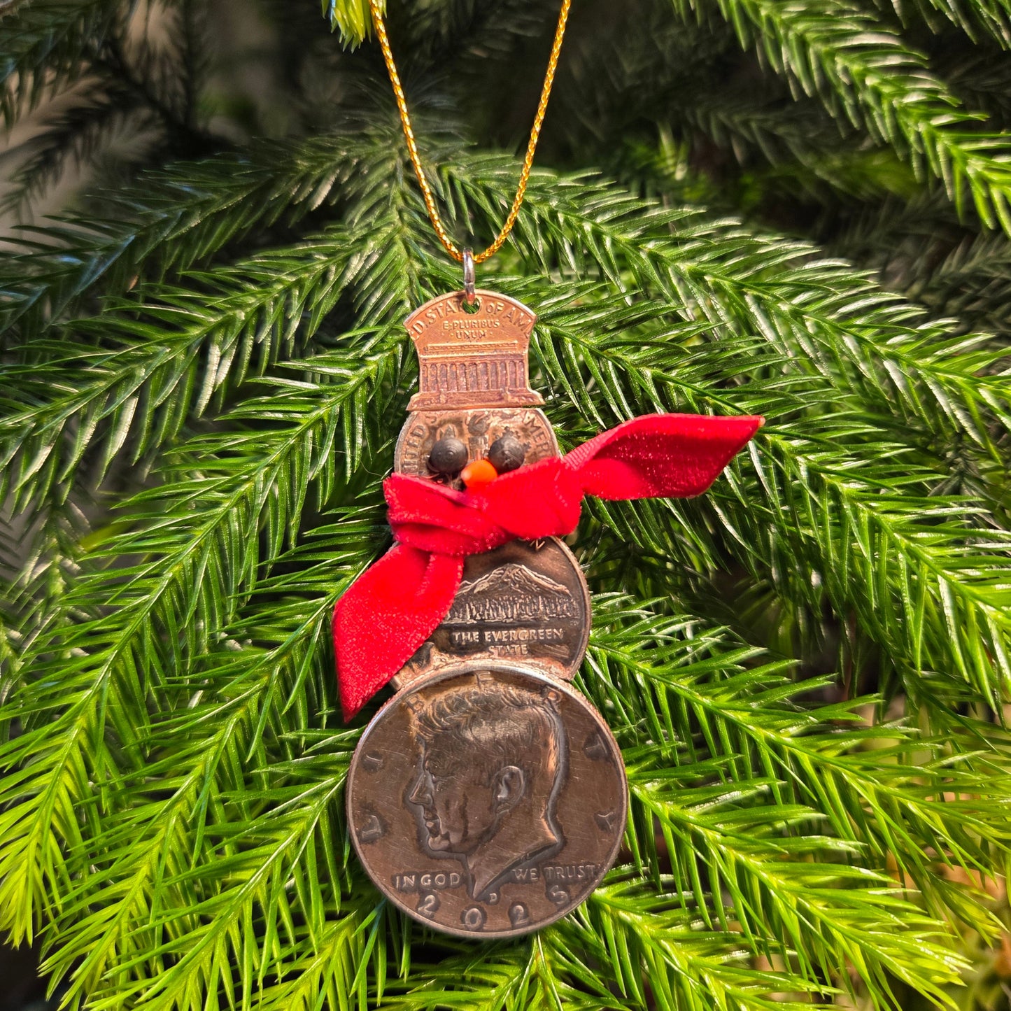 2025 Snowman Coin Christmas Ornament with a selection of U.S. coins (penny, dime, state quarter, and 2025 uncirculated half dollar) soldered into a snowman with a red ribbon scarf and golden tassel in front of a green pine tree. - Mountainstruck Coin Jewelry