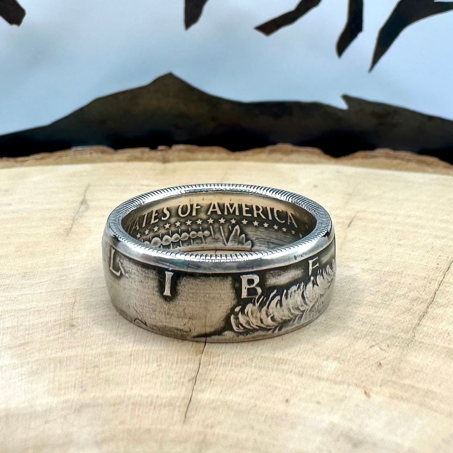U.S. JFK 1964 Half Dollar Silver Coin Ring on a light wood round with a metal mountain background. - Mountainstruck Coin Jewelry