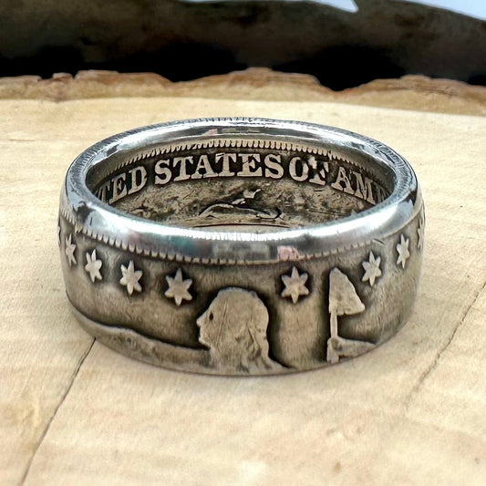 U.S. Seated Liberty Silver Half Dollar Coin Ring (patina) on a light wood round with a metal mountain background. - Mountainstruck Coin Jewelry