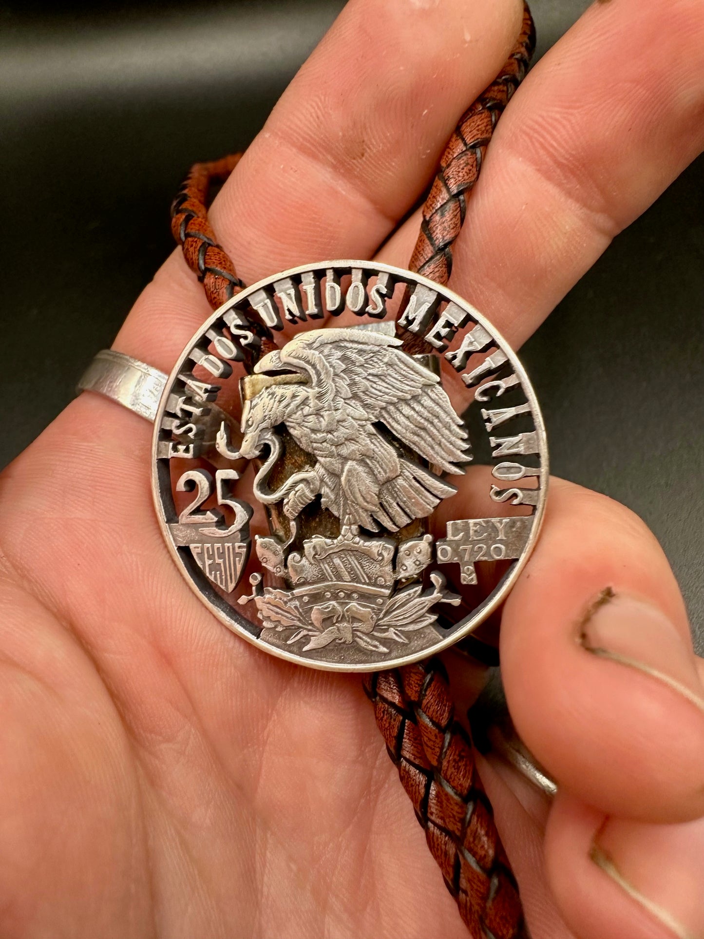 Mexico Silver 25 Pesos Hand Cut Coin Bolo Tie with darker brown leather cord on a hand for display.