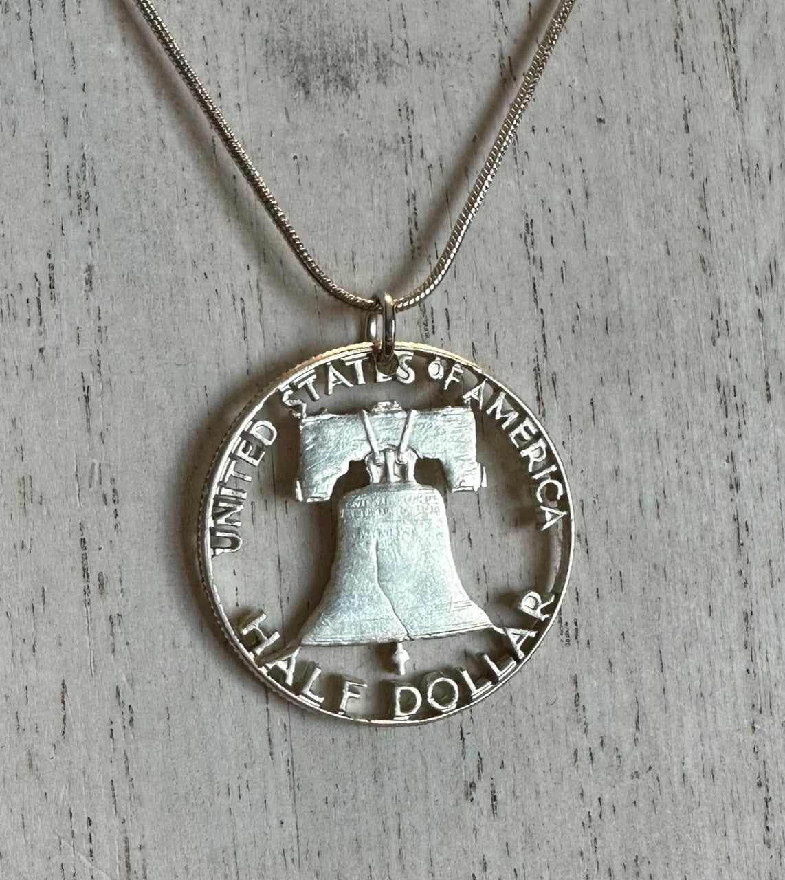 Cracked Liberty Bell U.S. Half Dollar Hand Cut Coin Necklace with snake chain on a white wood background.