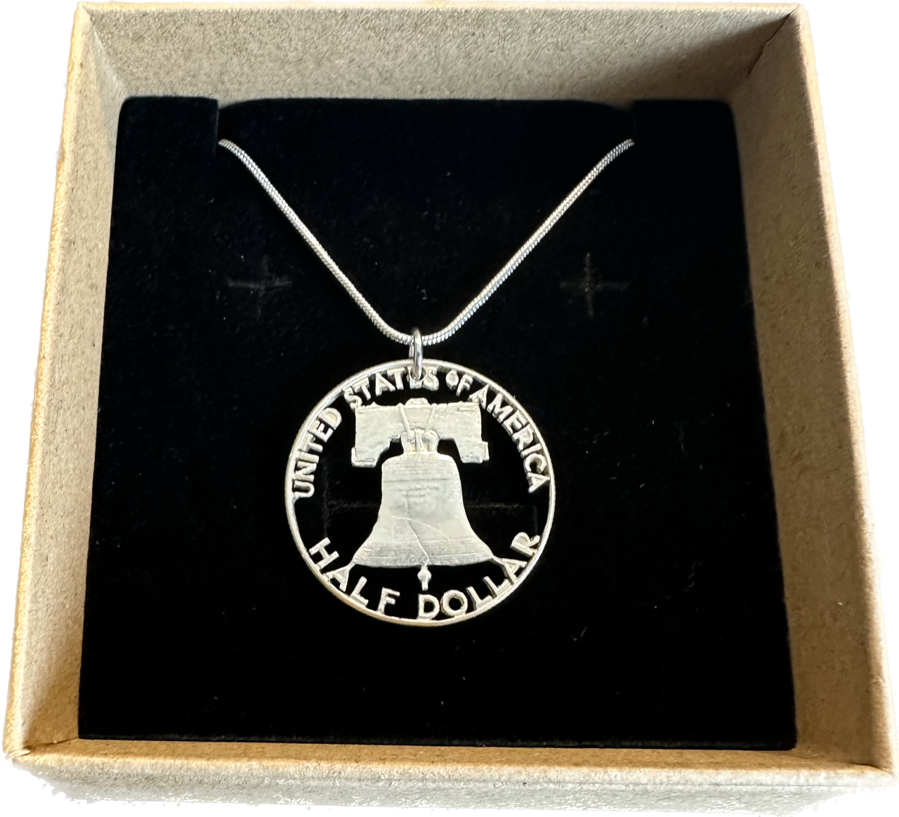 Cracked Liberty Bell U.S. Half Dollar Hand Cut Coin Necklace with a snake chain in a brown kraft box on a black velvet background.