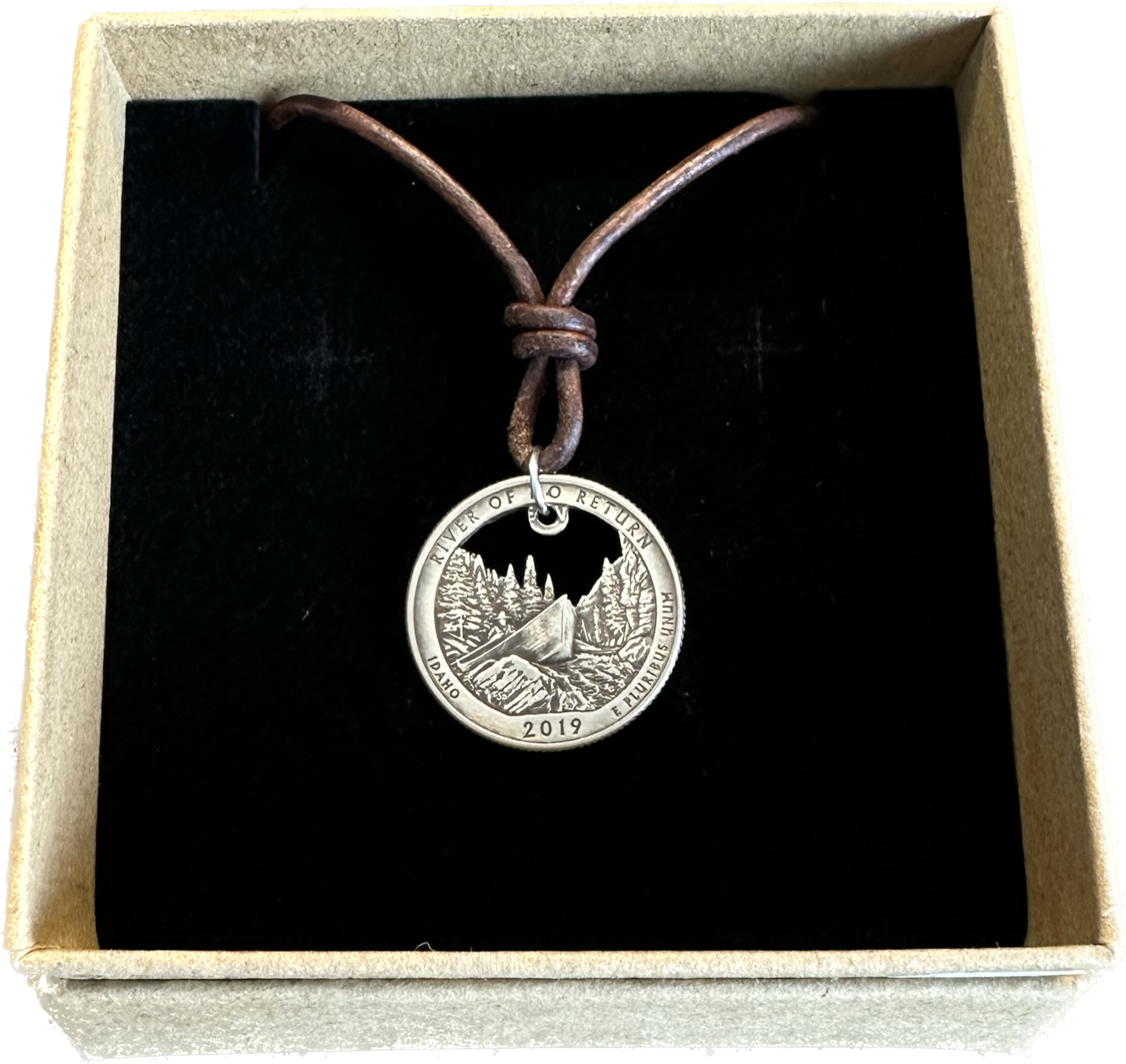 Idaho "River of No Return" Hand Cut Quarter Necklace with a leather cord in a brown kraft box on a black velvet background.