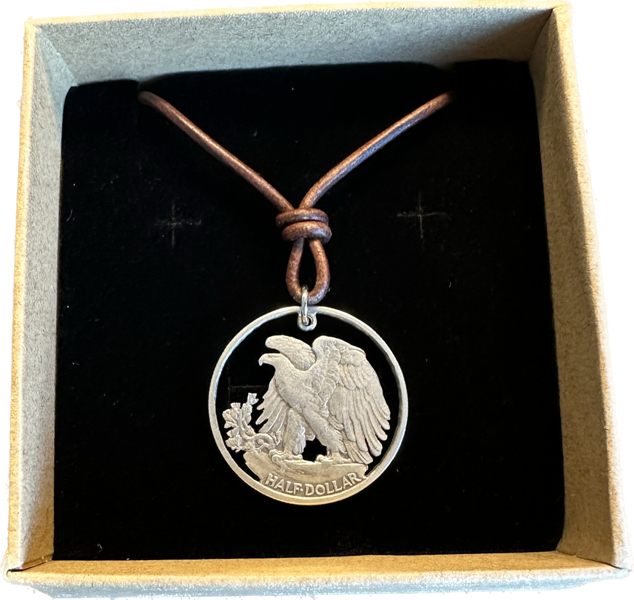 U.S. Walking Liberty Silver Hand Cut Coin Necklace (Eagle) with a leather cord in a brown kraft box on a black velvet background.