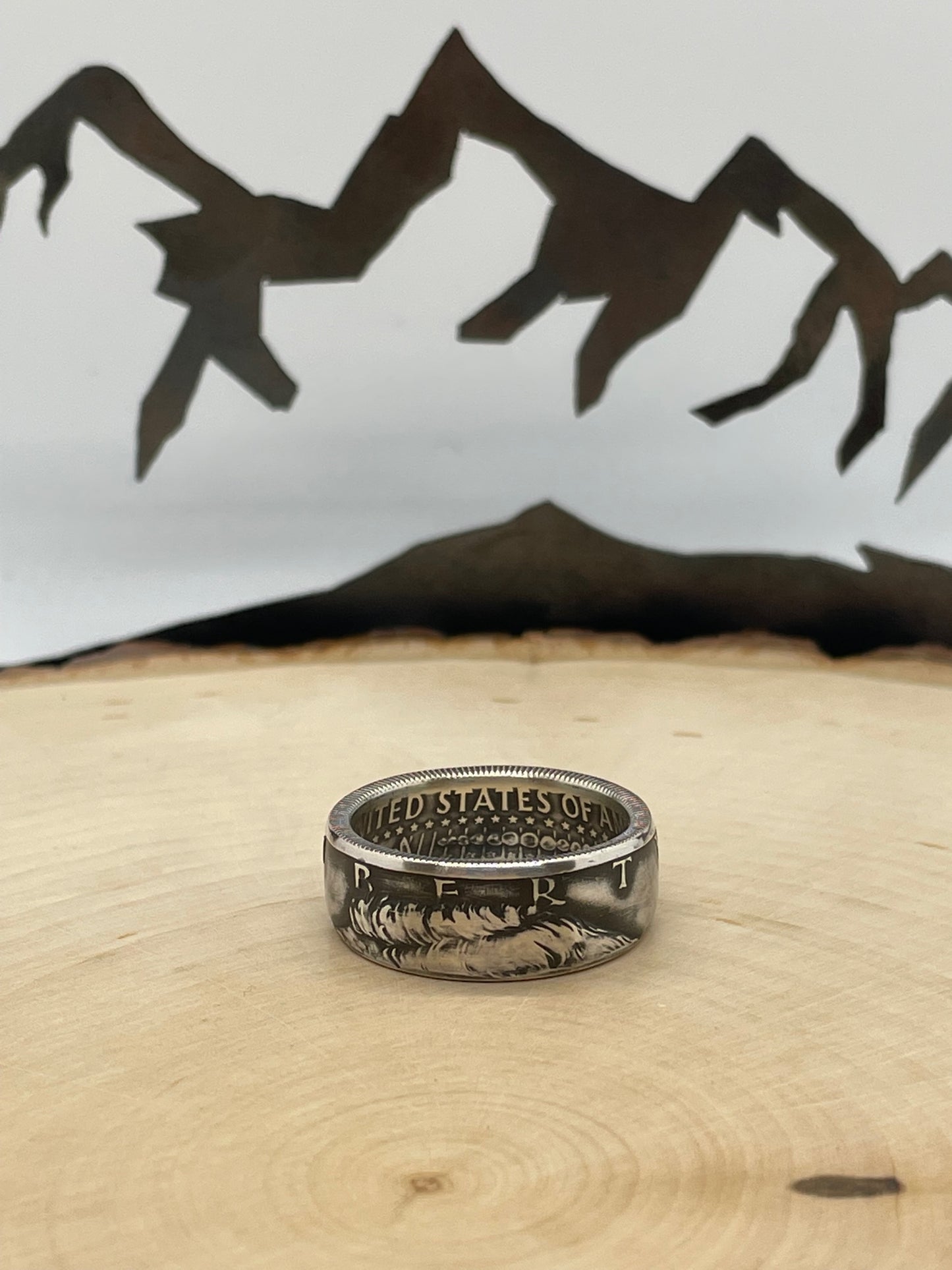 JFK Silver Half Dollar Coin Ring (front) on a light wood round with mountain outline in background.