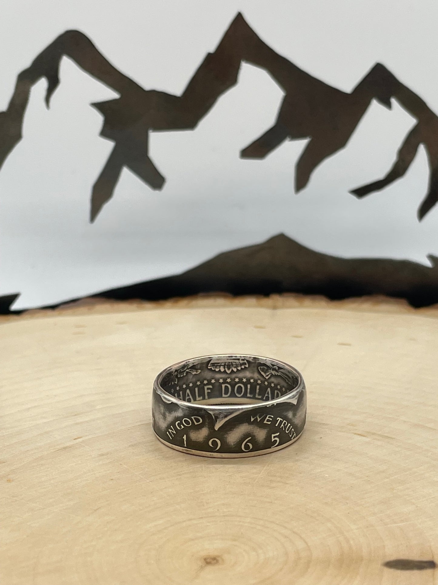 JFK Silver Half Dollar Coin Ring (front) on a light wood round with mountain outline in background.