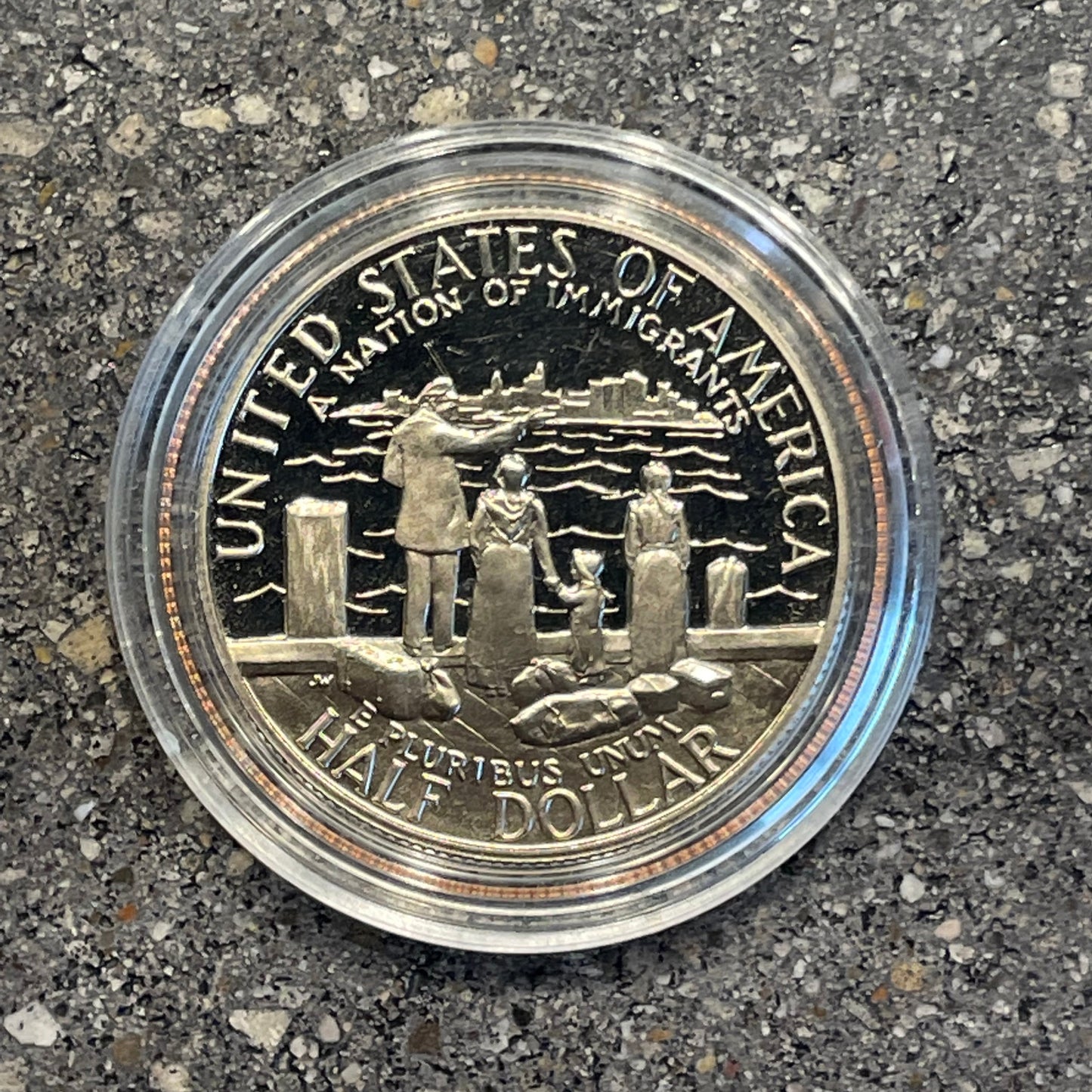 A Nation of Immigrants Half Dollar back of coin in a plastic case on a concrete background. - Mountainstruck Coin Jewelry