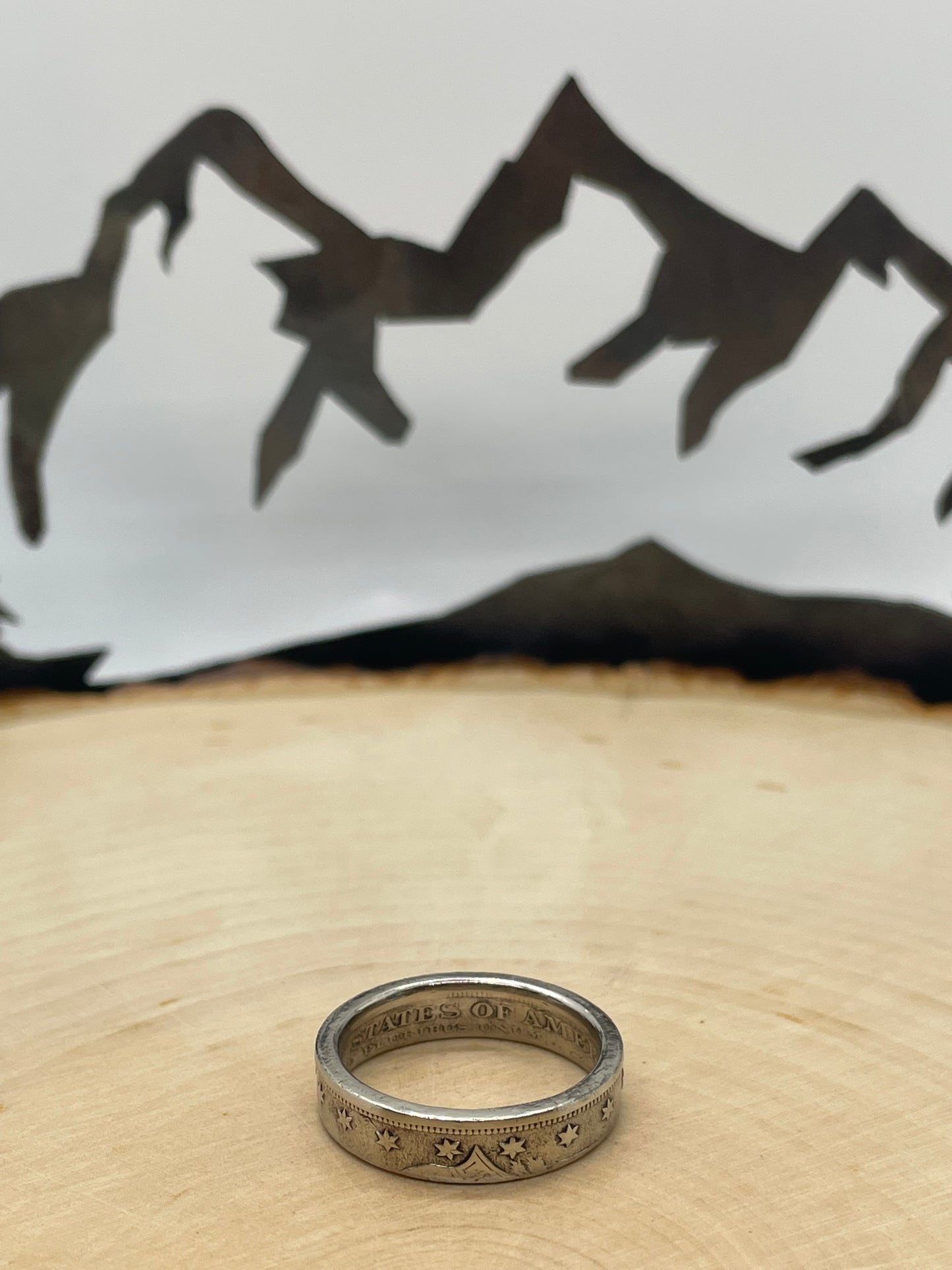 "V" Nickel Coin Ring on wood round with mountain outline in background.