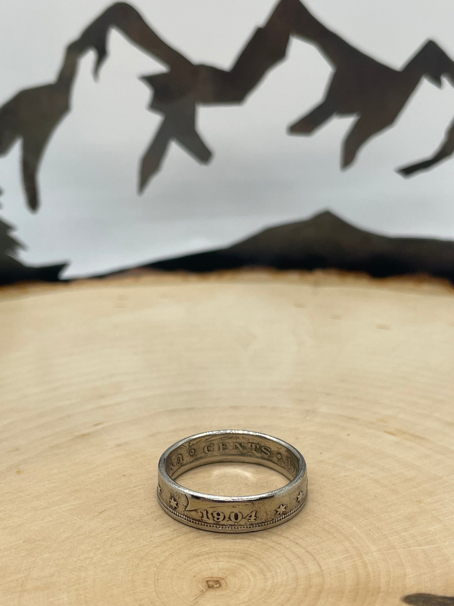 "V" Nickel Coin Ring on wood round with mountain outline in background.