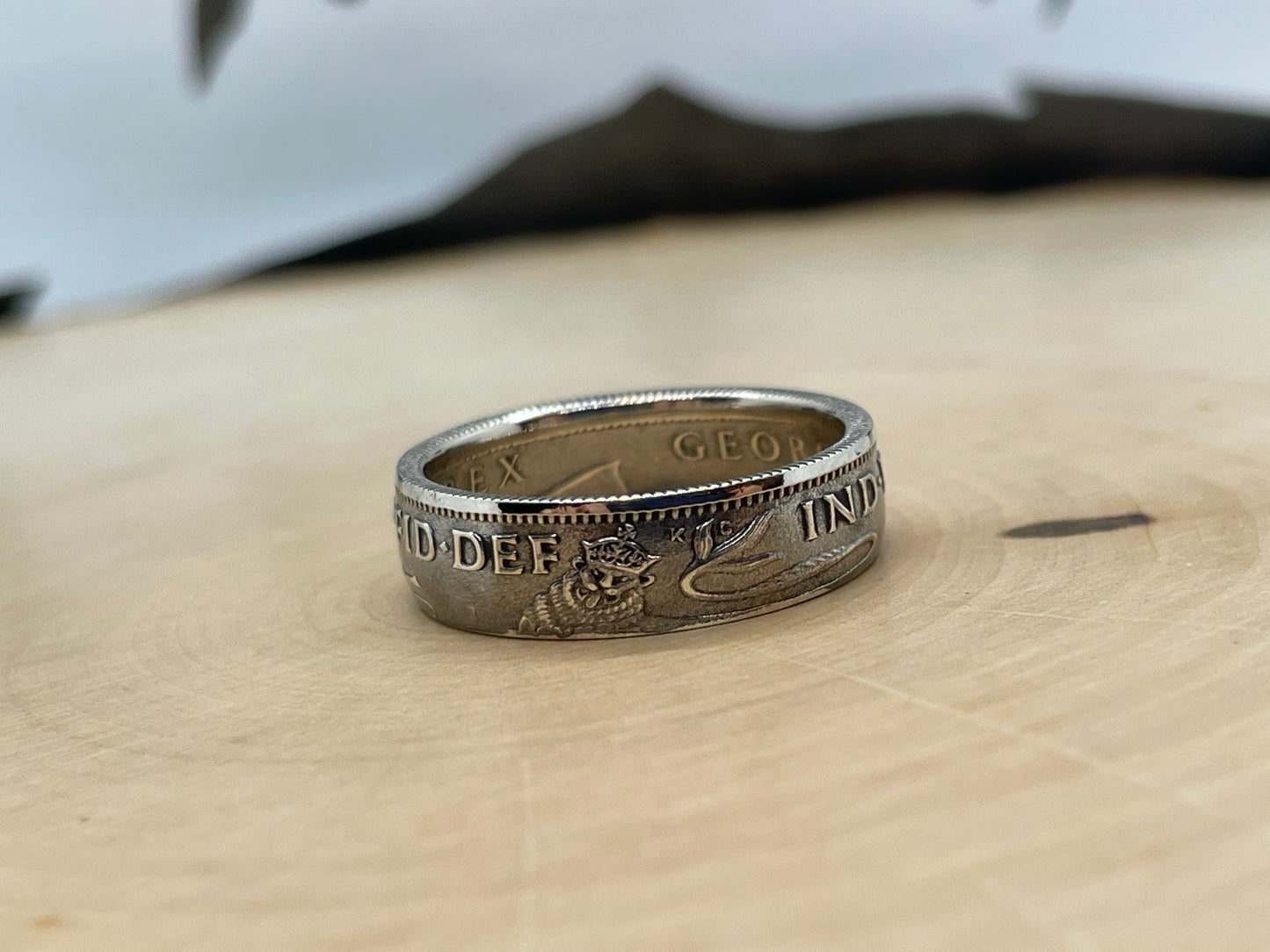 UK One Shilling Silver Coin Ring on a light wood round with a mountain outline in the background.