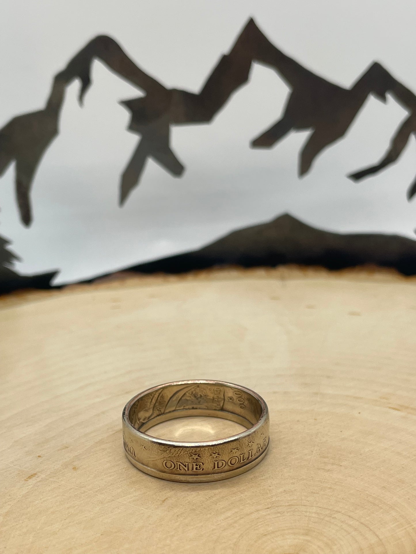 Sacagawea Dollar Coin Ring on a light wood round with mountain outline in background.