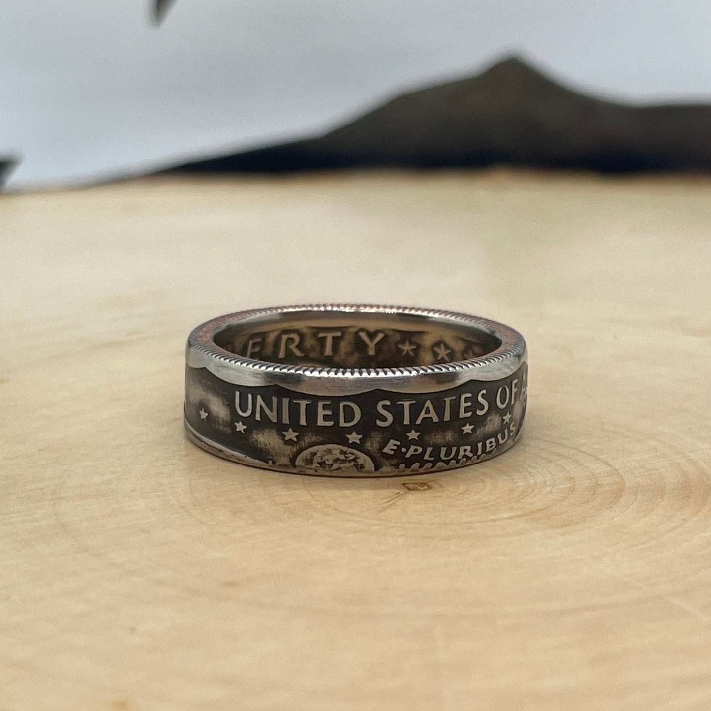Susan B. Anthony Dollar Coin Ring on a light wood round with mountain outline in background.
