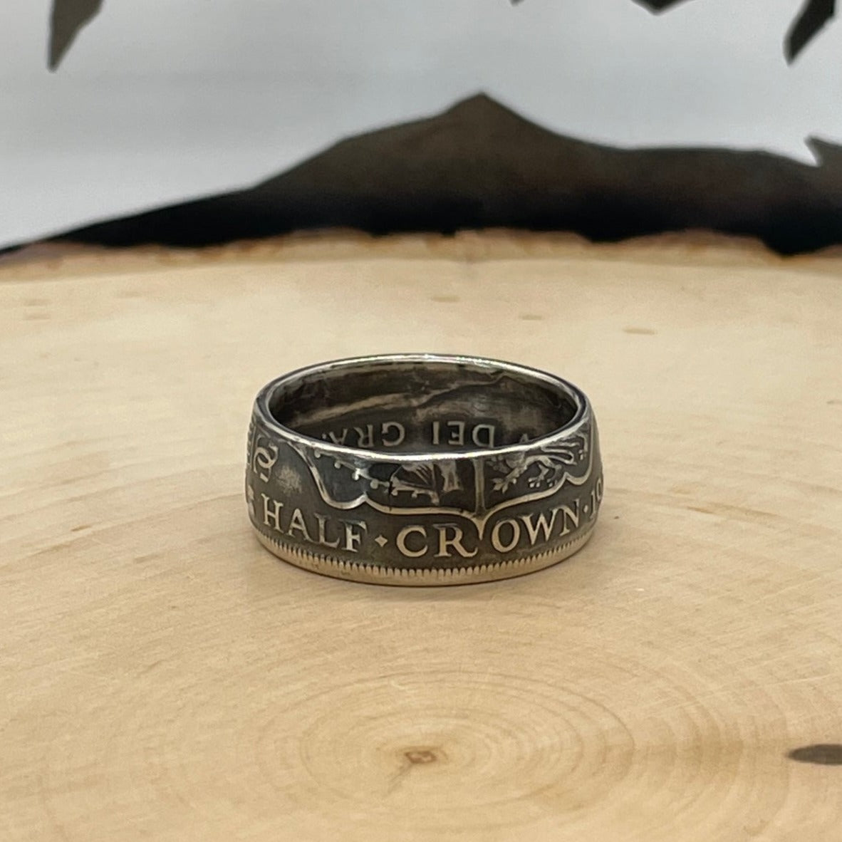 UK Half Crown Silver Coin Ring (1927-1936, G.B.) on a light wood round with mountain outline in background.