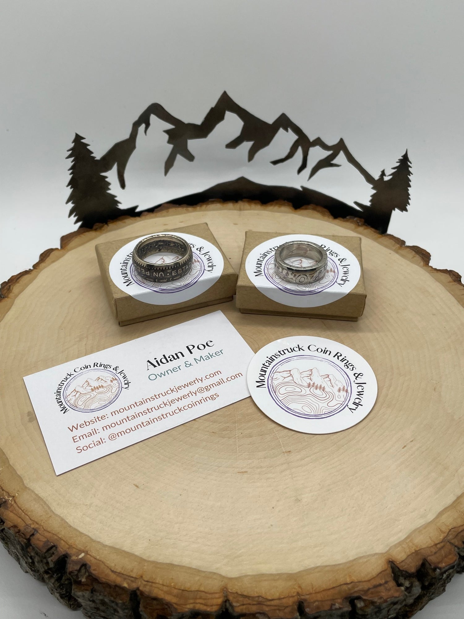 Mountainstruck Coin Rings & Jewelry business card, sticker and two small ring boxes on a thing tree stump with a metal art tree and mountain design in the background. There is a ring sitting on top of each of the boxes.