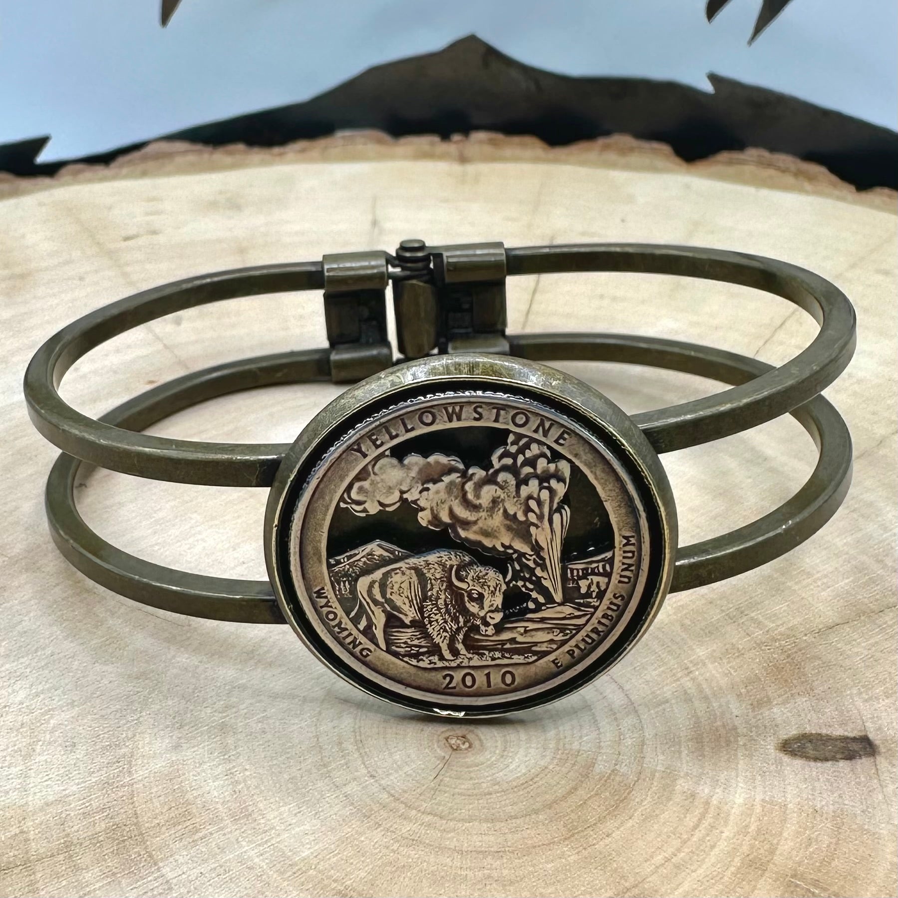 U.S. National Park Quarter Hand Cut Coin Bronze Bracelet on a light wood round with a metal mountain background. - Mountainstruck Coin Jewelry