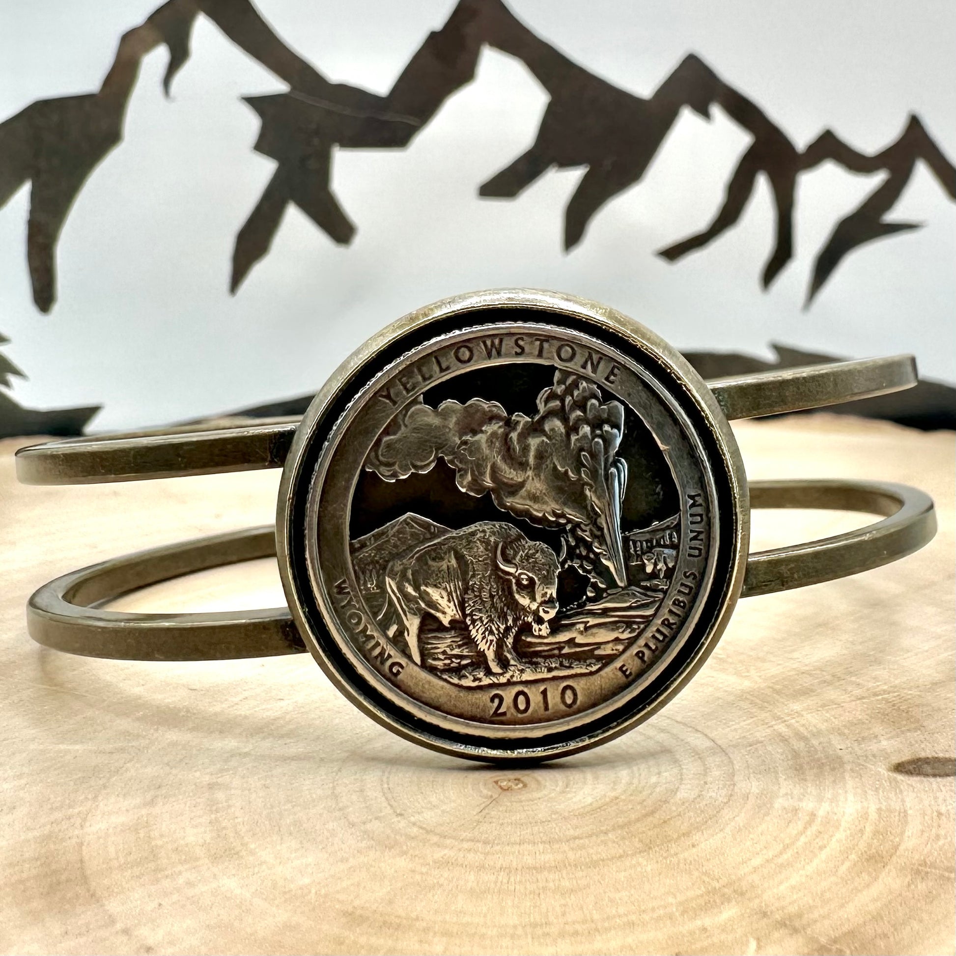U.S. National Park Quarter Hand Cut Coin Bronze Bracelet on a light wood round with a metal mountain background. - Mountainstruck Coin Jewelry