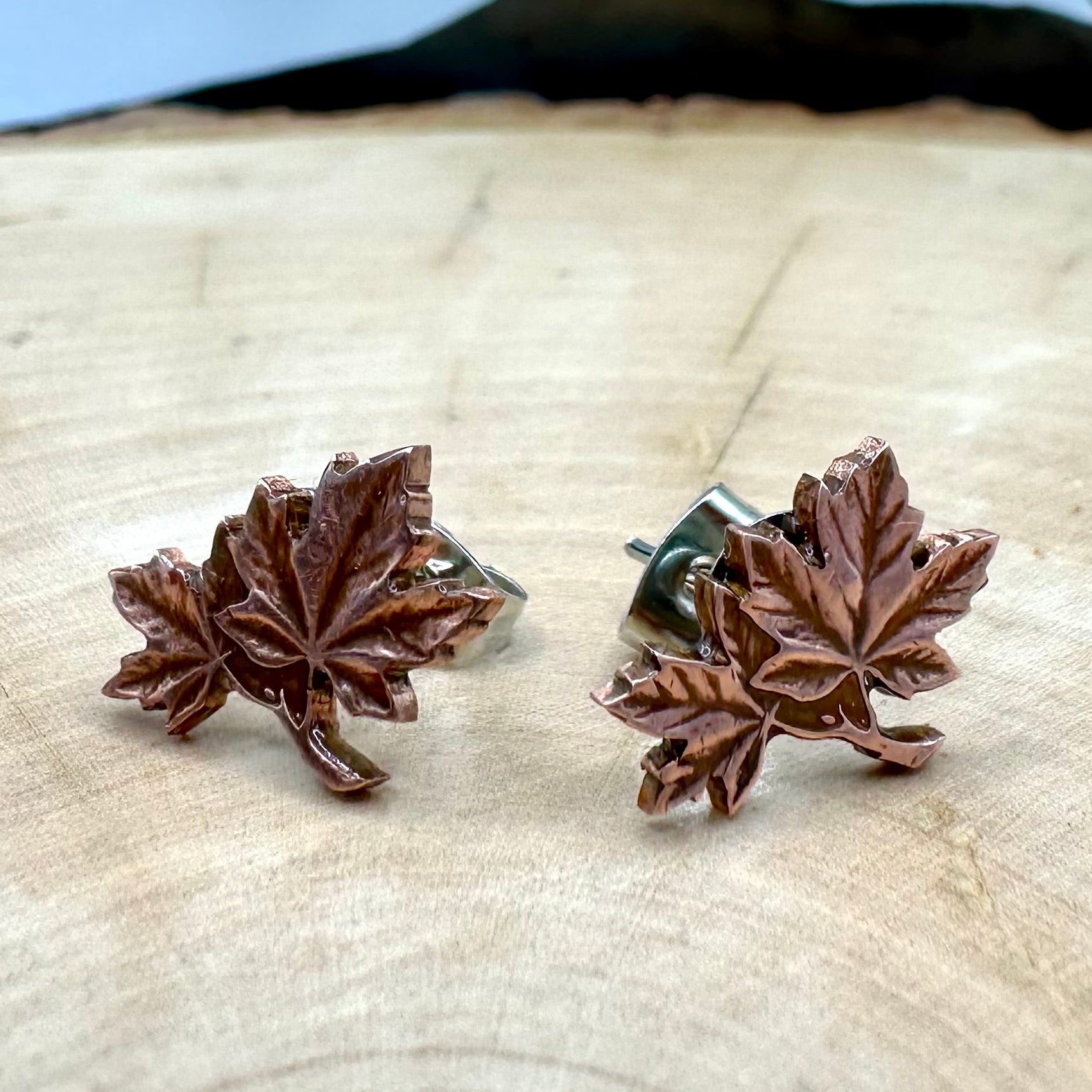 Canada Maple Leaf Penny Stud Coin Earrings on a light wood round metal mountain background. 
- Mountainstruck Coin Jewelry
