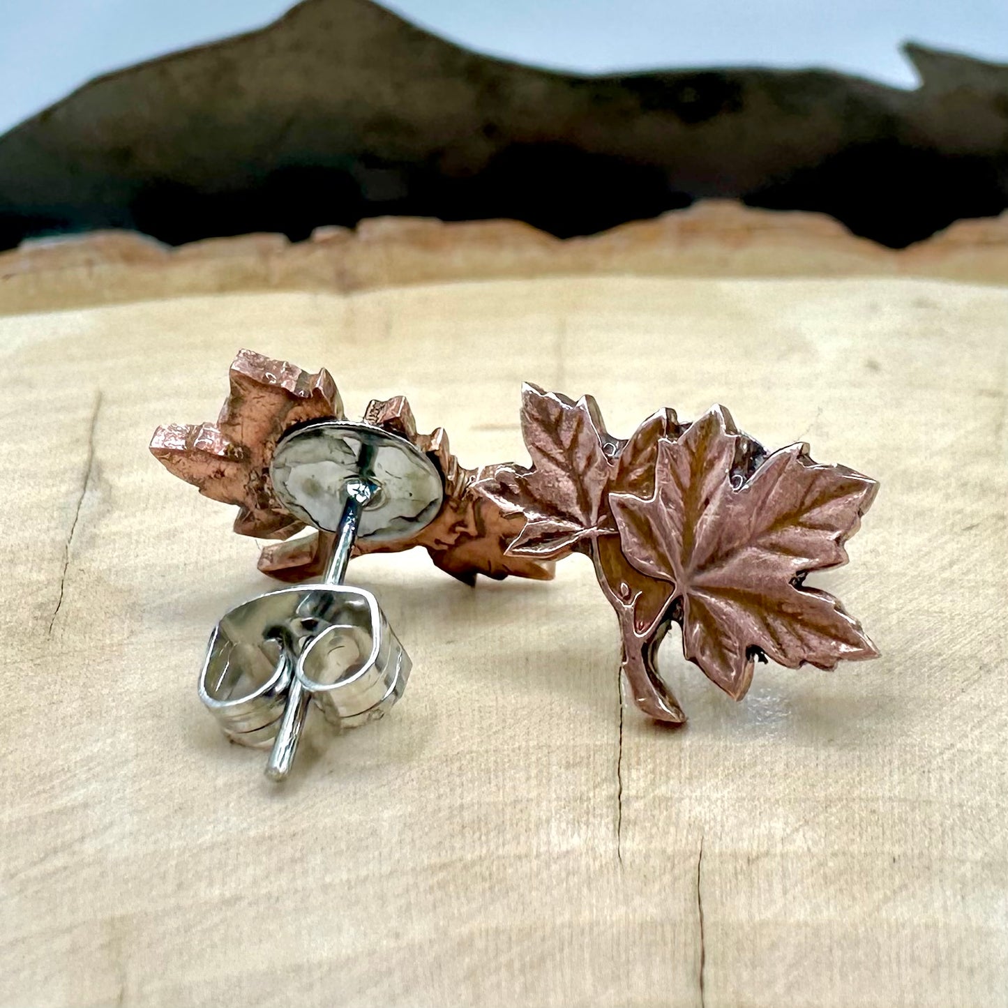Canada Maple Leaf Penny Stud Coin Earrings on a light wood round metal mountain background. 
- Mountainstruck Coin Jewelry