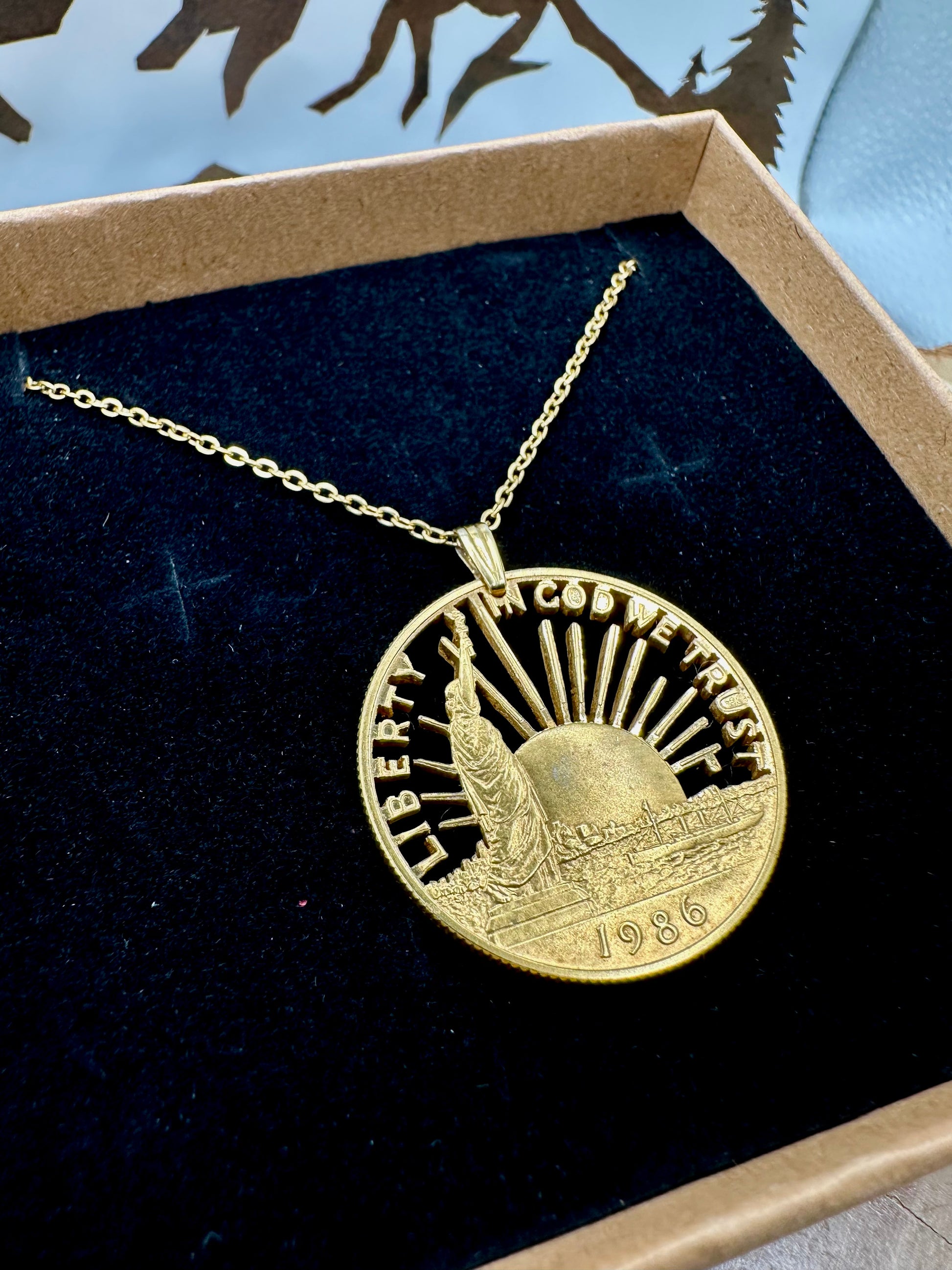 24k Plate U.S. Nation of Immigrants Cut Half Dollar Coin Necklace in a brown kraft box on a black velvet background with a metal mountain display. - Mountainstruck Coin Jewelry