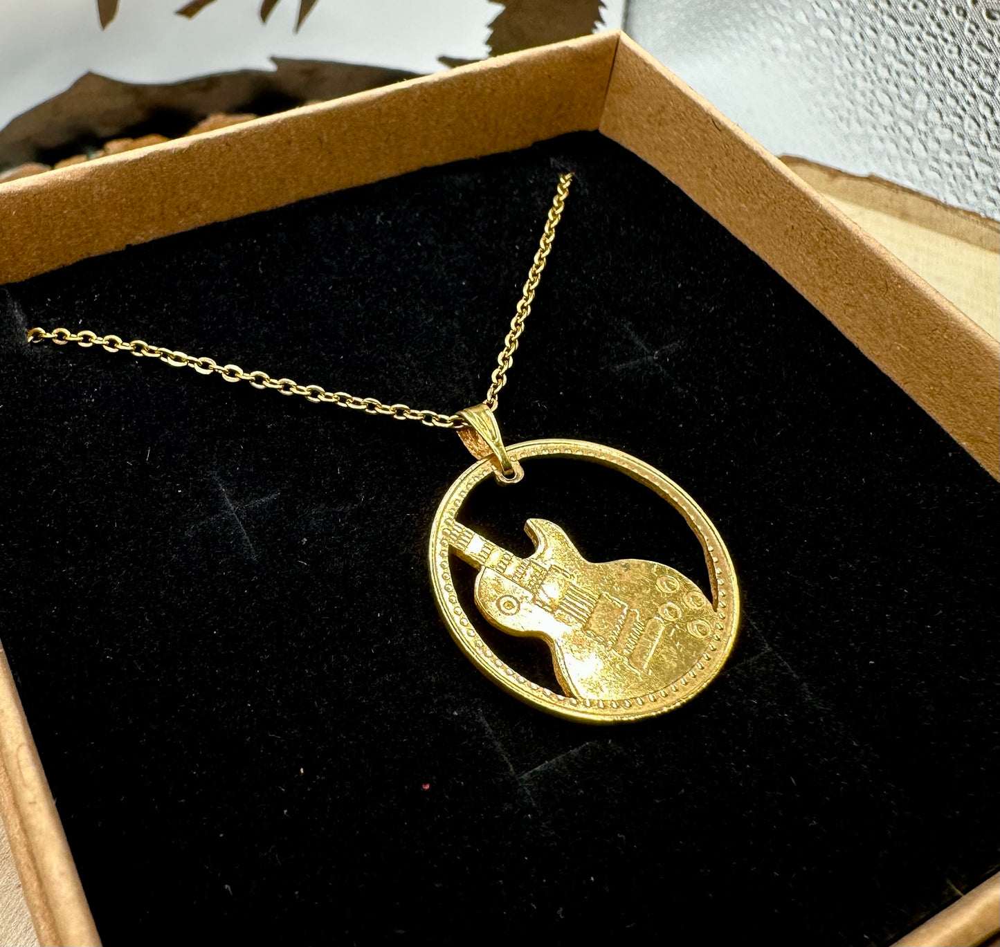 Guitar Hand Cut Token Necklace in a brown kraft box on a black velvet background on top of a light wood round with a metal mountain display. - Mountainstruck Coin Jewelry