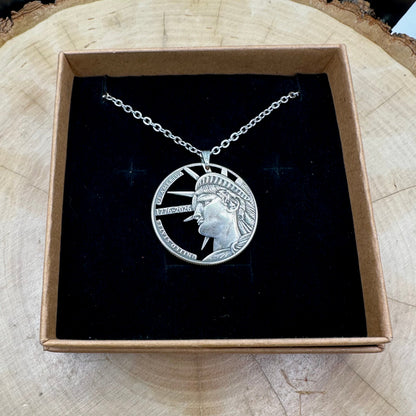 U.S. Half Dollar Hand Cut Coin Necklace (front) with a stainless chain on a black velvet background in a brown kraft box on a wood round. - Mountainstruck Coin Jewelry