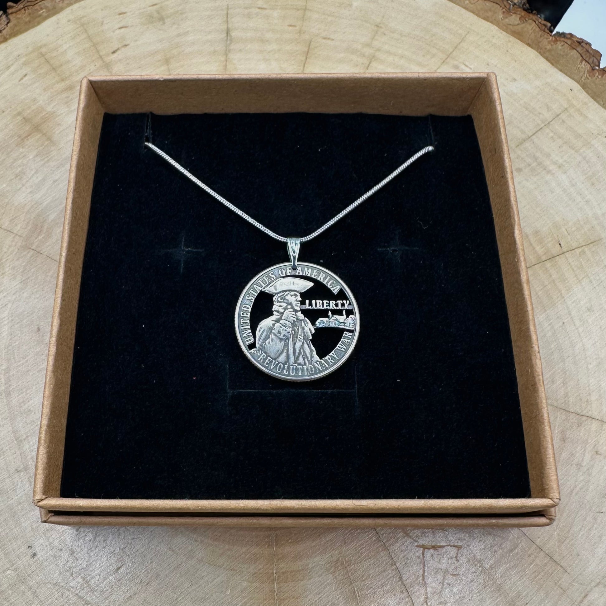 U.S. 2026 "Revolutionary War" Quarter Hand Cut Coin Necklace with a snake chain on a black velvet background in a brown kraft box on a wood round. - Mountainstruck Coin Jewelry
