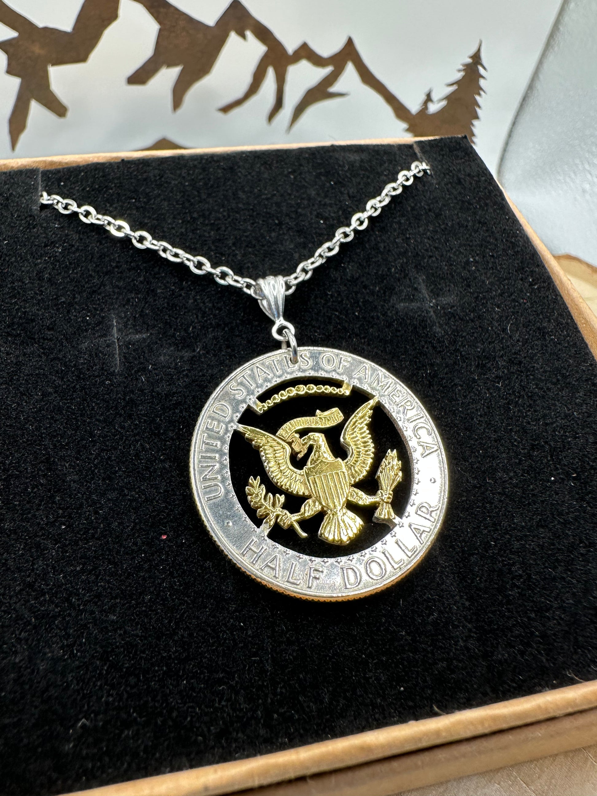 U.S. JFK Eagle Half Dollar Hand Cut Silver Coin Necklace with 24k gold plated Eagle in a brown kraft box on a black velvet background with a metal mountain display. - Mountainstruck Coin Jewelry