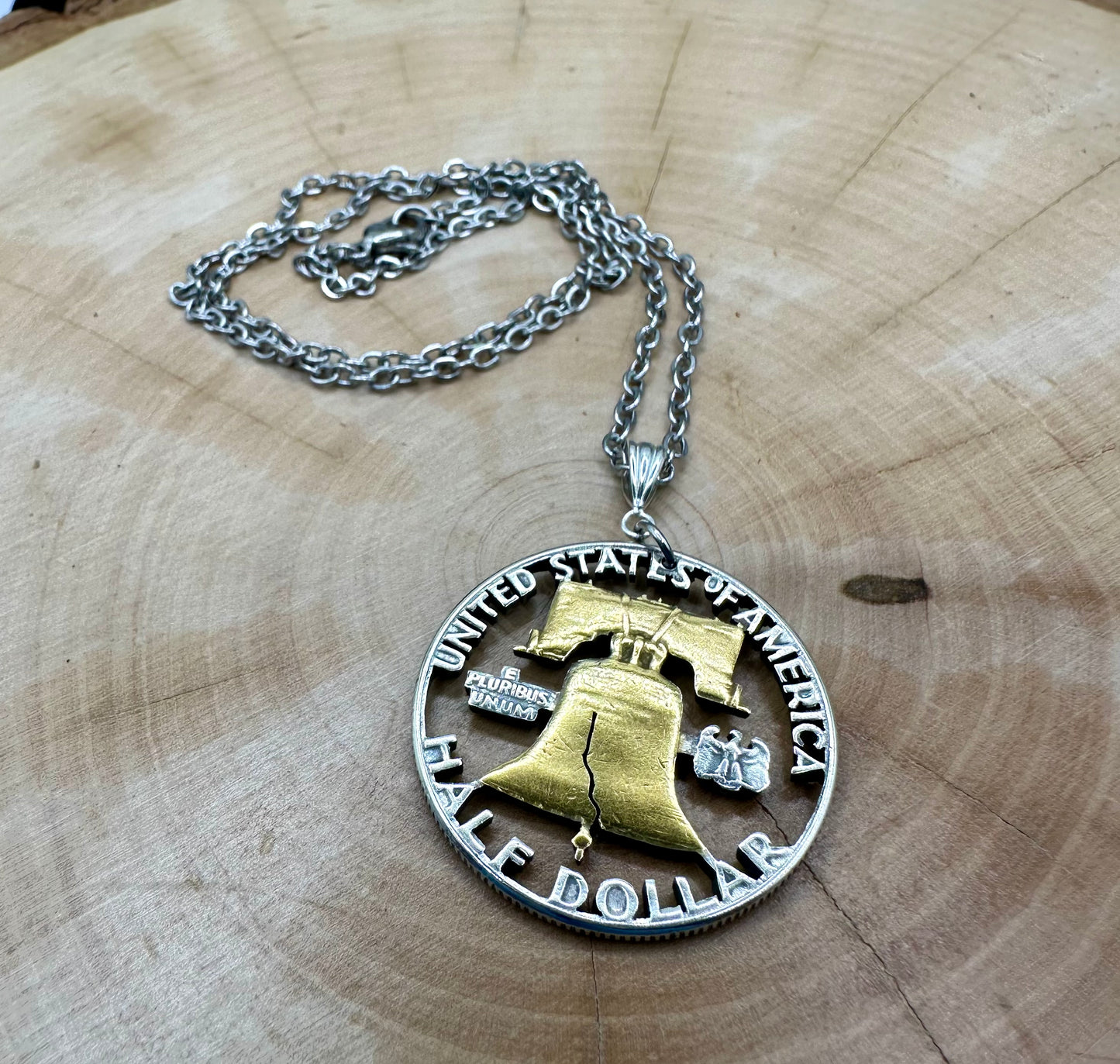 U.S. Cracked Liberty Bell Half Dollar Hand Cut Coin Necklace 24k Gold Plated Bell with a stainless chain on a light wood background. - Mountainstruck Coin Jewelry
