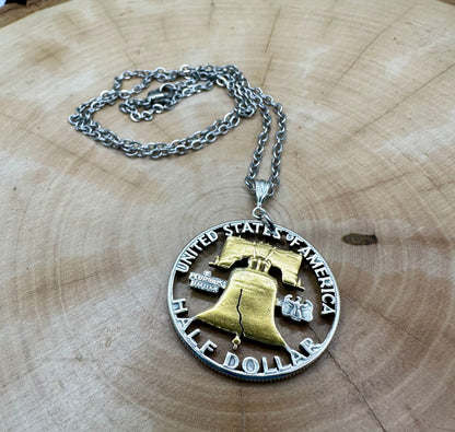 U.S. Cracked Liberty Bell Half Dollar Hand Cut Coin Necklace 24k Gold Plated Bell with a stainless chain on a light wood background. - Mountainstruck Coin Jewelry