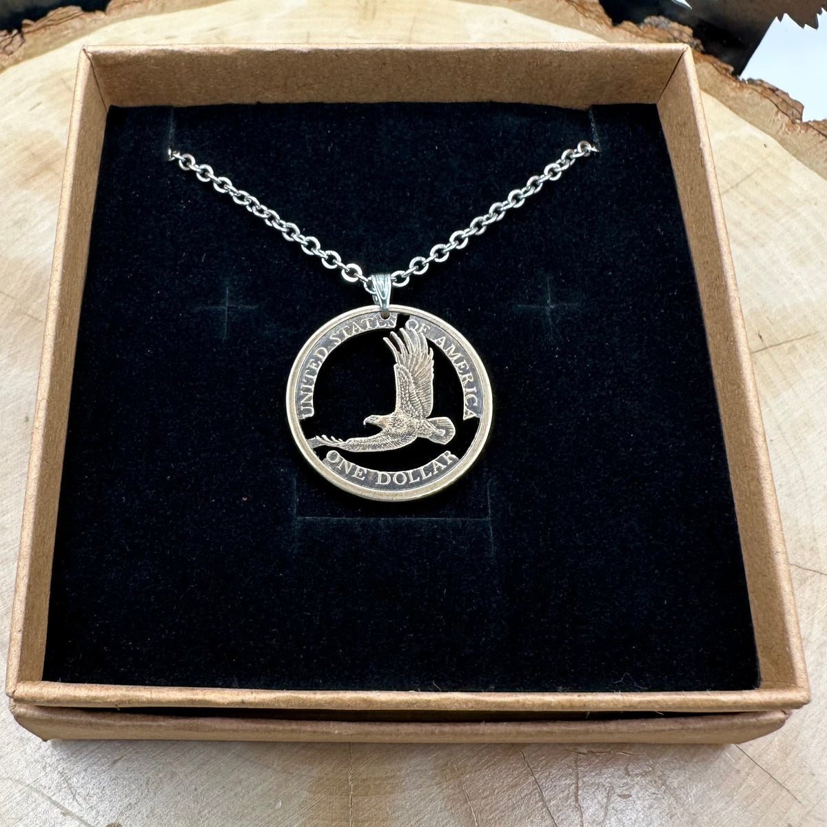 U.S. Flying Eagle Sacajawea One Dollar Hand Cut Coin Necklace with snake chain in a brown kraft box on a black velvet background on top of a light wood round with metal mountain and tree display. - Mountainstruck Coin Jewelry