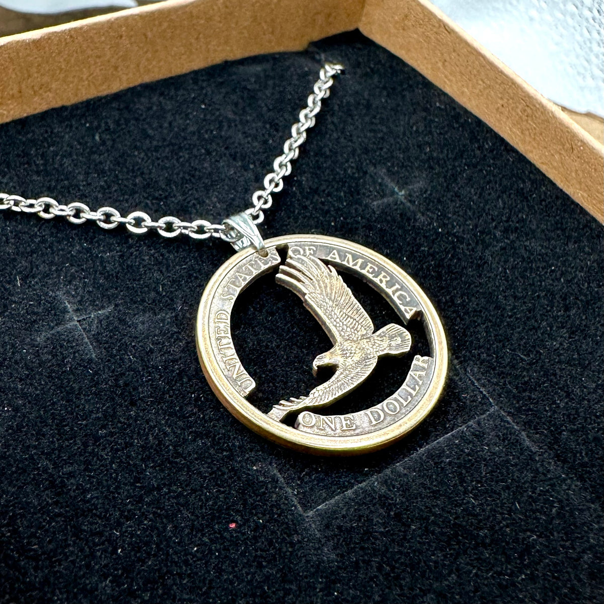 U.S. Flying Eagle Sacajawea One Dollar Hand Cut Coin Necklace with snake chain in a brown kraft box on a black velvet background. - Mountainstruck Coin Jewelry