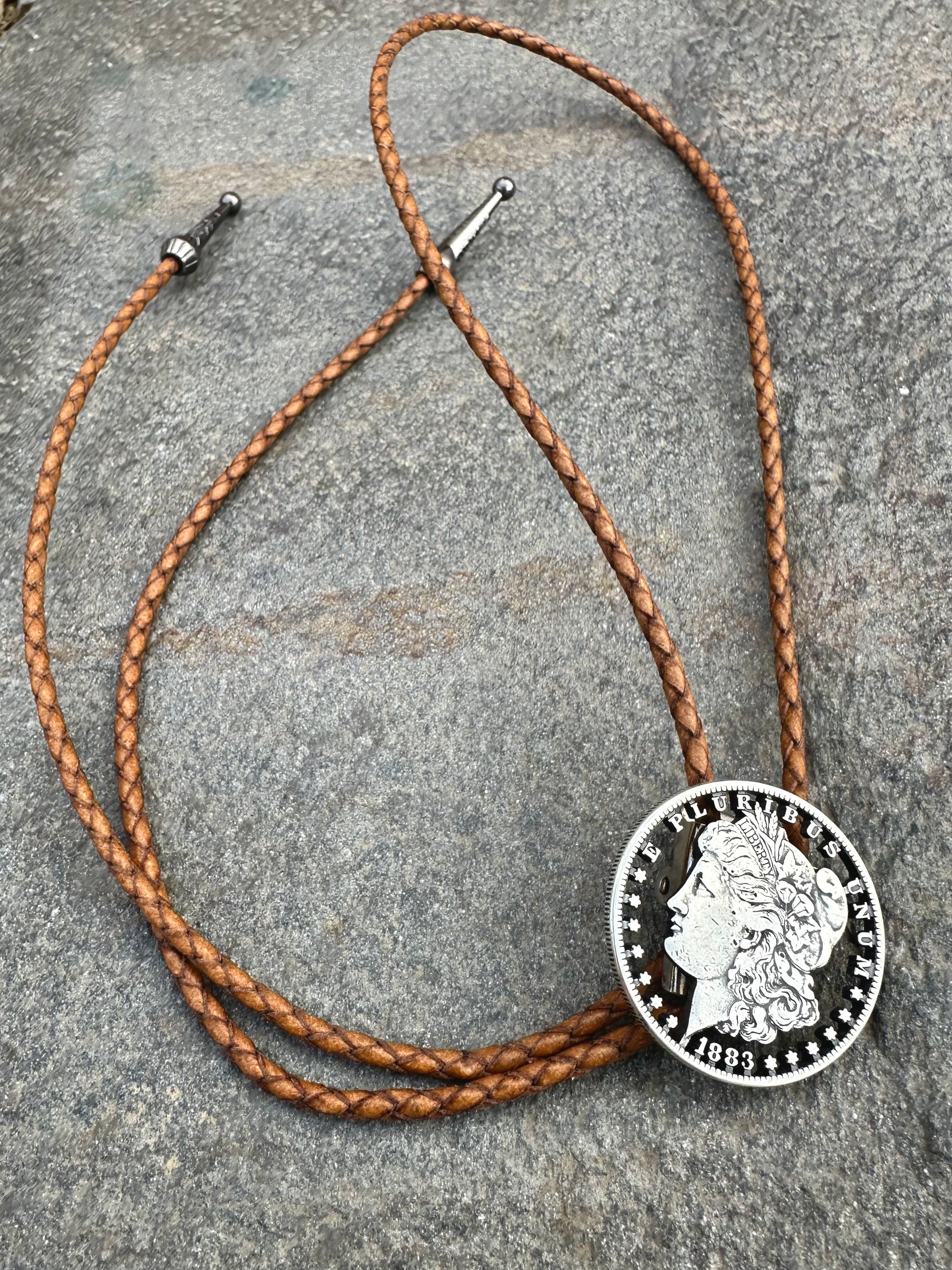 U.S. Morgan Hand Cut Silver Dollar Coin Bolo Tie (front) on a grey rock surface. - Mountainstruck Coin Jewelry