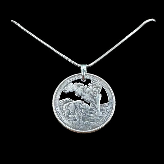 U.S. Yellowstone National Park Quarter Hand Cut Coin Necklace in a brown kraft box on a black velvet background on top of a light wood round with a metal mountain and tree behind. - Mountainstruck Coin Jewelry