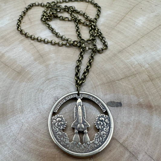 American Innovation $1 Florida Space Shuttle Hand Cut Coin necklace on a light wood background. - Mountainstruck Coin Jewelry