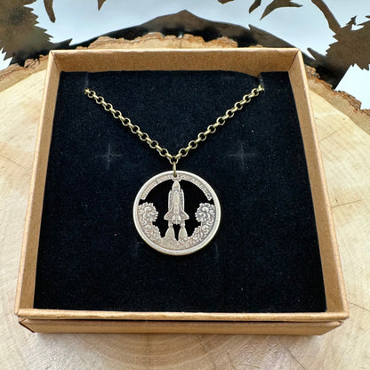American Innovation $1 Florida Space Shuttle Hand Cut Coin necklace in a brown kraft box on a black velvet background on top of a light wood round with metal mountains and trees in the background. - Mountainstruck Coin Jewelry