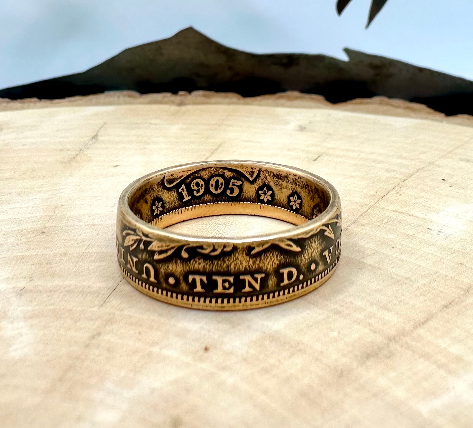 22k U.S. Gold Eagle $10 Coin Ring on a light wood round with a metal mountain background. - Mountainstruck Coin Jewelry