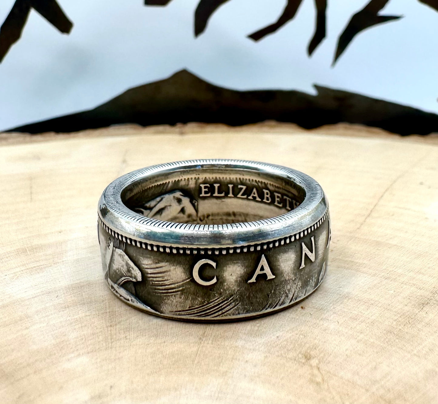Canada Dollar Silver Coin Ring on a light wood round with a metal mountain background. - Mountainstruck Coin Jewelry