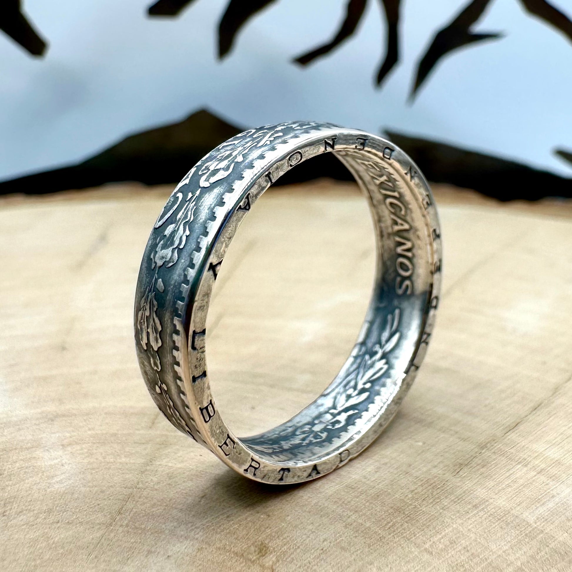 Mexico 50 Centavos Silver Coin Ring (Rare) on its edge on a light wood round with mountain outline in the background. 
- Mountainstruck Coin Jewelry