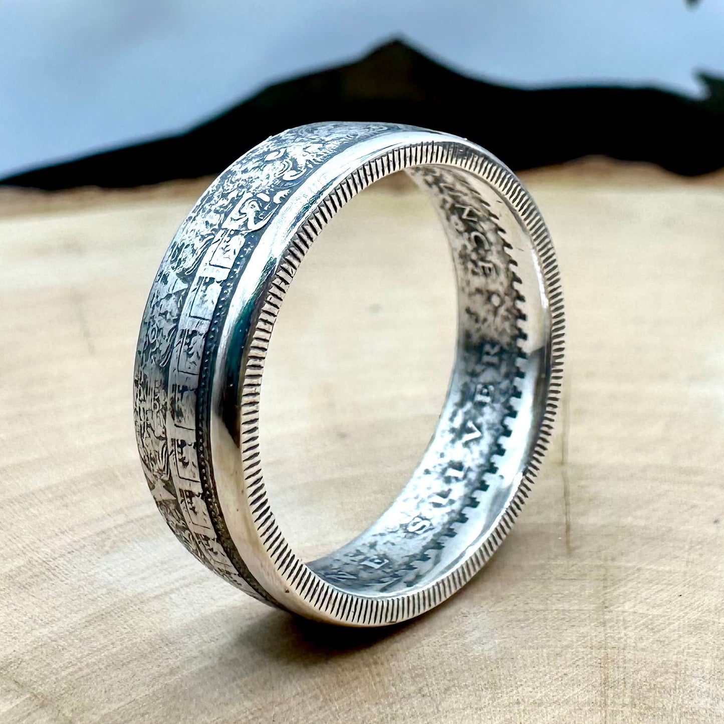 Mexico Aztec Calendar 1/4 oz Silver Round Ring on edge on a light wood round with a metal mountain background. 
- Mountainstruck Coin Jewelry