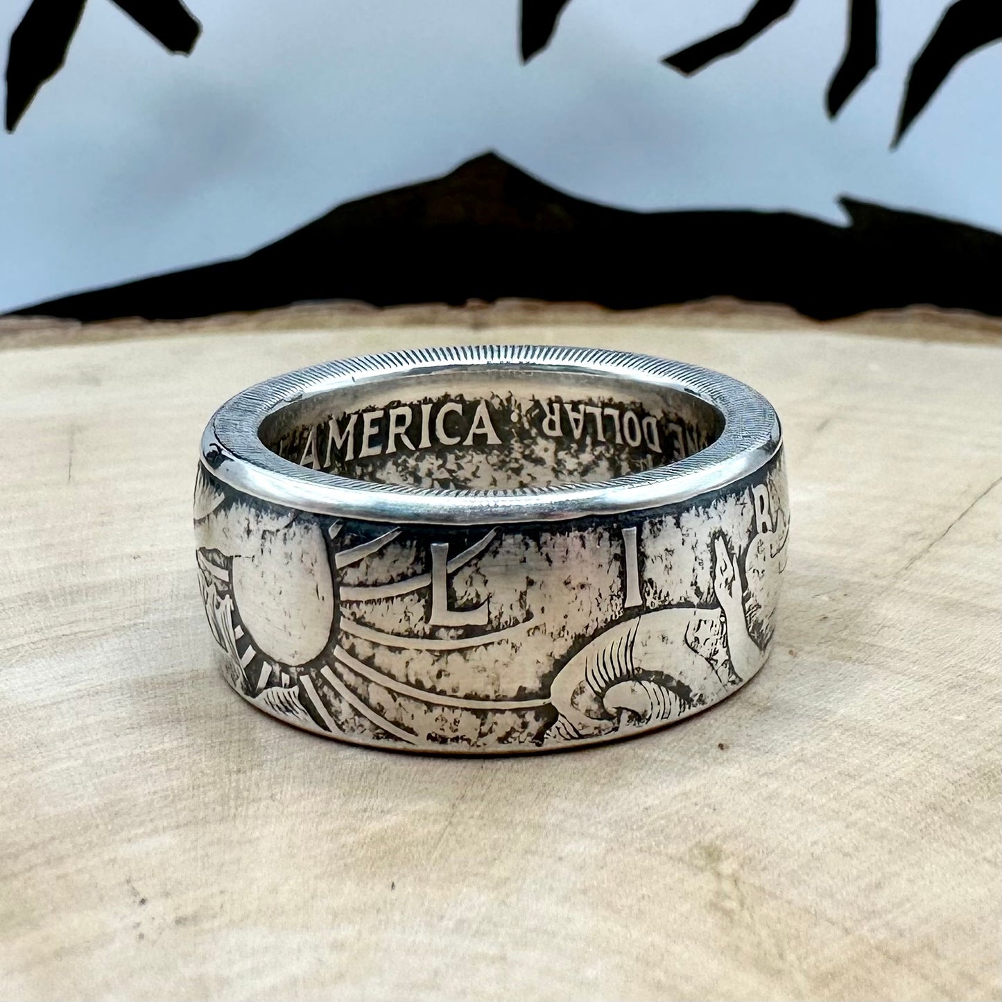 U.S. 1oz 999 Silver Eagle $1 Coin Ring on a light wood round with a metal mountain background. - Mountainstruck Coin Jewelry