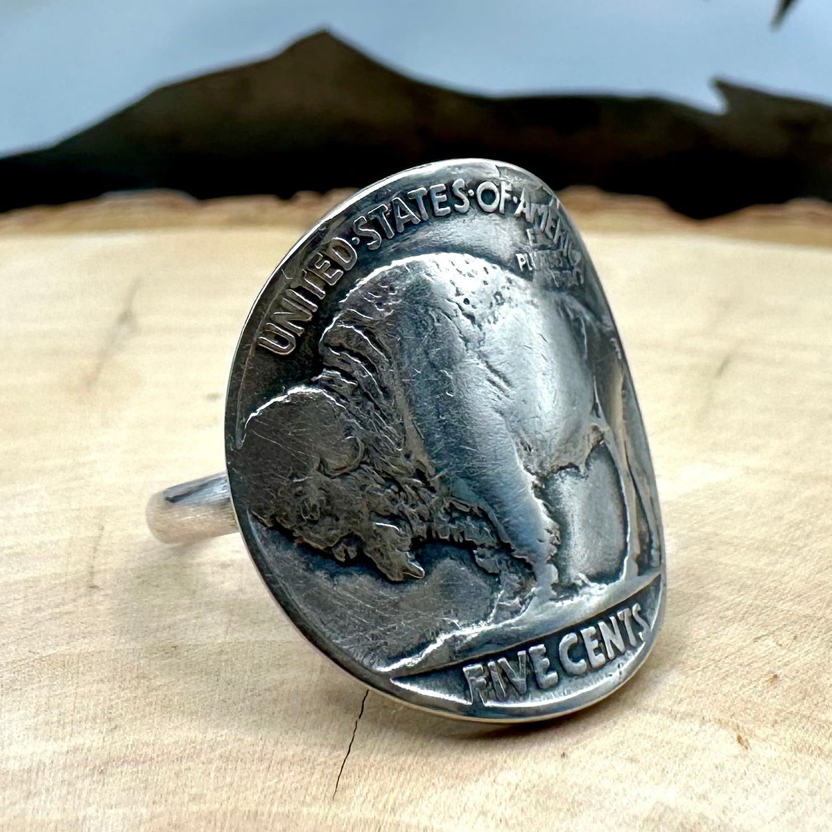 U.S. Buffalo Nickel Coin Signet Ring (Buffalo Side) with a sterling silver band on a light wood round with a metal mountain background. - Mountainstruck Coin Jewelry