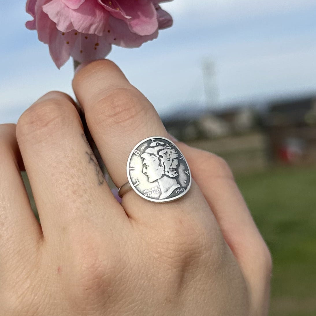 Hand wearing a U.S. Mercury Dime Signet Ring holding a pink flower with a blurred outdoor background.- Mountainstruck Coin Jewelry 