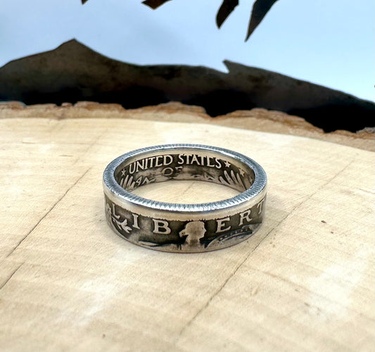 U.S. Standing Liberty Quarter Silver Coin Ring ion a light wood round with a metal mountain background. - Mountainstruck Coin Jewelry