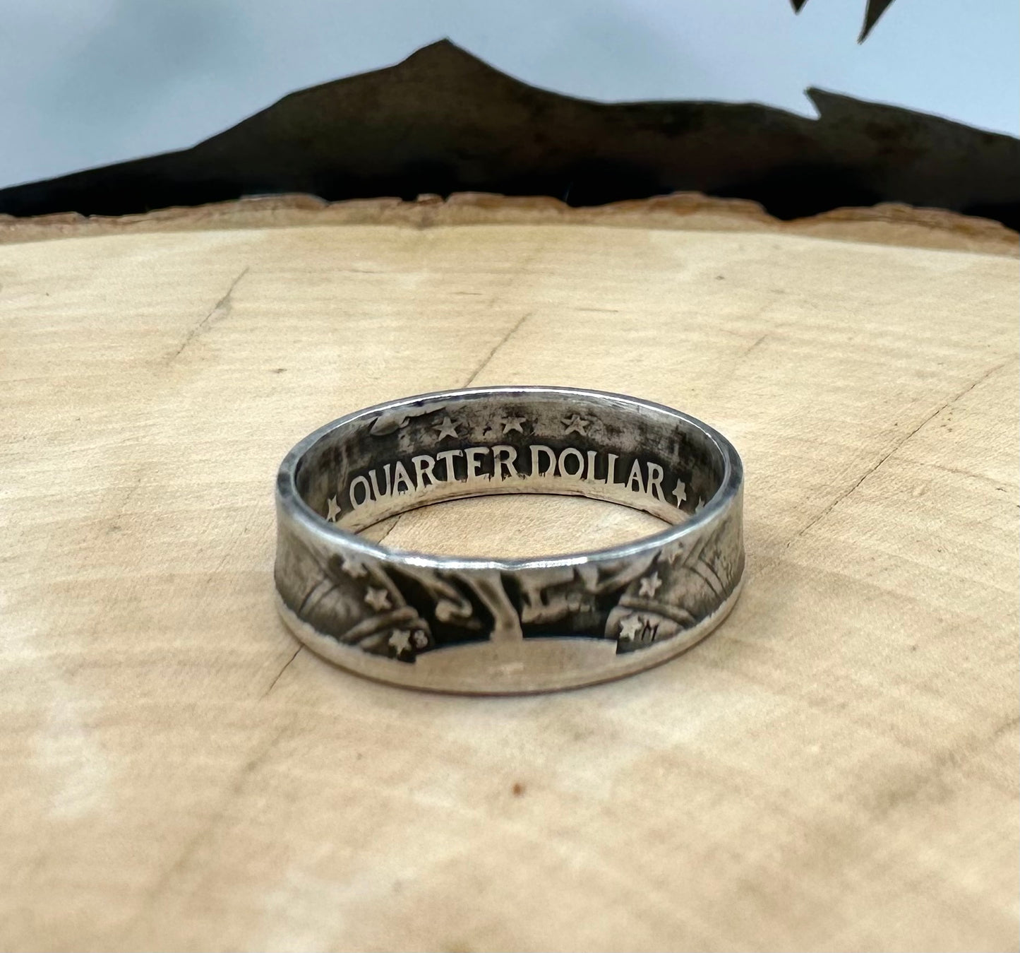 U.S. Standing Liberty Quarter Silver Coin Ring ion a light wood round with a metal mountain background. - Mountainstruck Coin Jewelry